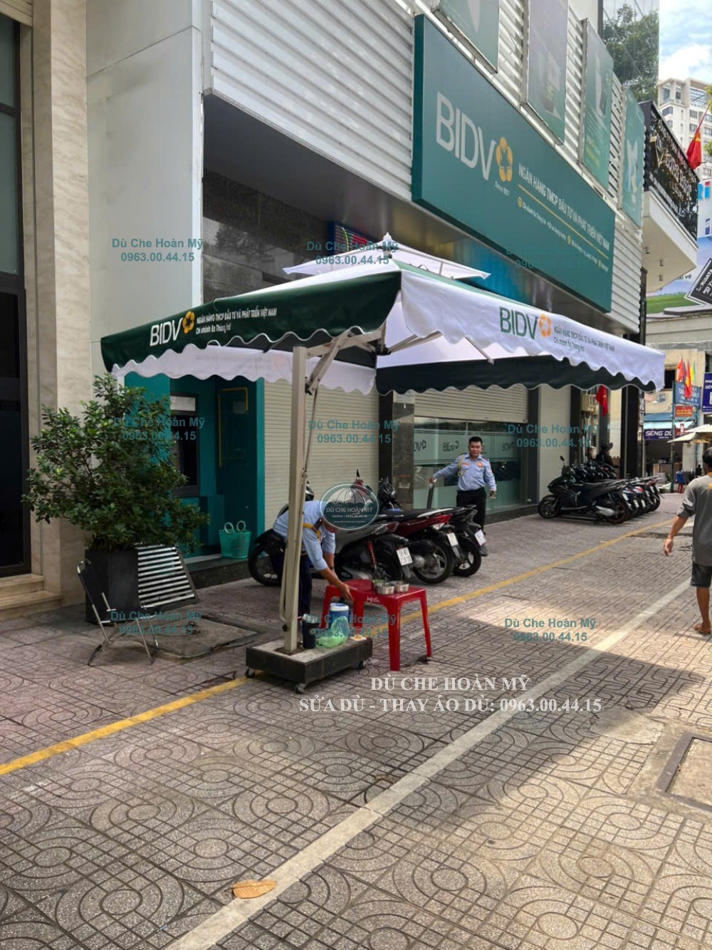 Dù vuông lệch tâm xanh–trắng được bố trí phía trước BIDV, với cột đặt sát vùng bên ngoài tán.